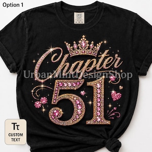 May include: A black short-sleeved t-shirt featuring a graphic design with the text Chapter 51 in gold and pink jewel-toned lettering. A crown sits above the text, and the number 51 is decorated with pink gems. Small heart and star accents surround the text.