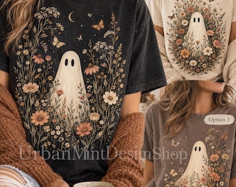 May include: Three t-shirts in black, cream, and brown, each featuring a graphic of a ghost surrounded by wildflowers, butterflies, and a crescent moon. The text UrbanMintDesignShop is visible on the black shirt. Each shirt has a crew neck.