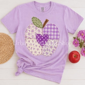 May include: A light purple short-sleeve t-shirt featuring a large apple-shaped applique on the front. The apple is divided into sections of green gingham, purple floral, and purple gingham fabric, with a central purple heart and a brown stem.