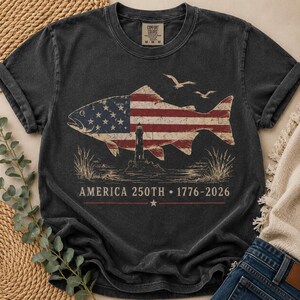 May include: A charcoal grey short-sleeve t-shirt featuring a graphic of a fish filled with the American flag pattern, a lighthouse, and two birds. Below the graphic, the text reads AMERICA 250TH 1776-2026 with a small star centred underneath.