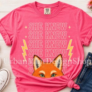 May include: A bright pink short-sleeved t-shirt featuring the repeated white text SHE KNEW in a retro font, flanked by two yellow lightning bolts. Below the text is a graphic illustration of a fox face with orange fur and green eyes.