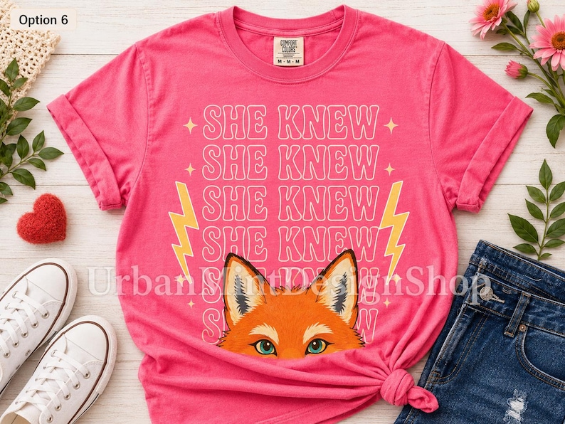 May include: A bright pink short-sleeved t-shirt featuring the repeated white text SHE KNEW in a retro font, flanked by two yellow lightning bolts. Below the text is a graphic illustration of a fox face with orange fur and green eyes.