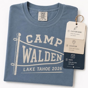 May include: A blue t-shirt featuring a white graphic of a pennant flag with the text CAMP WALDEN LAKE TAHOE 2026. Attached is a three-part tag with icons and text for custom name, location, and date, including instructions to enter the desired details.