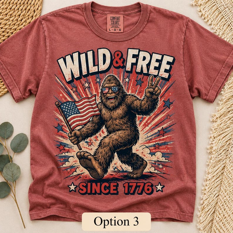 Puede incluir: Camiseta roja con un gr&aacute;fico de Bigfoot con gafas de sol y una bandera estadounidense. El texto "WILD & FREE" y "SINCE 1776" est&aacute;n impresos en la camiseta. La camiseta es de la marca Comfort Colors.