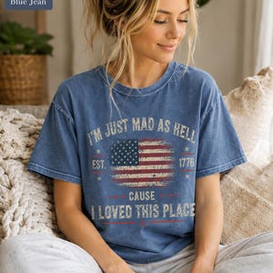 May include: Blue jean colored t-shirt with the text "I'M JUST MAD AS HELL" above a distressed American flag graphic. Below the flag, the text reads "CAUSE I LOVED THIS PLACE" and "EST. 1776".