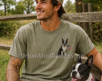 May include: An olive green crew neck t-shirt featuring a small graphic on the left chest of a black and white Boston Terrier dog sitting next to a small American flag. The shirt is made of a soft, washed cotton fabric with a classic fit.