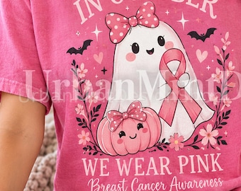 May include: A pink t-shirt featuring a graphic of a white ghost and a pink pumpkin, both wearing polka-dot bows. The ghost holds a pink ribbon. The text reads: IN OCTOBER WE WEAR PINK Breast Cancer Awareness. The design includes bats and flowers.