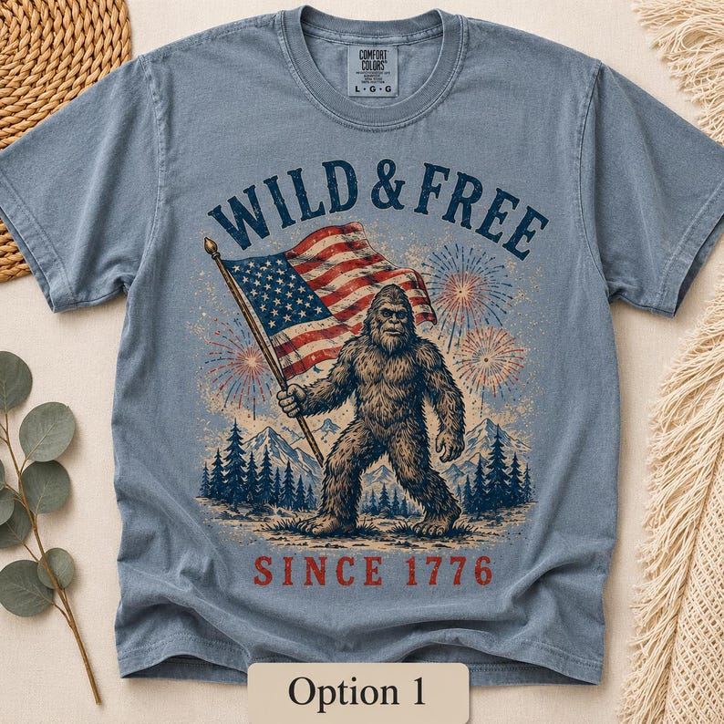 Puede incluir: Camiseta azul claro de manga corta con un dise&ntilde;o gr&aacute;fico que muestra a un Bigfoot sosteniendo una bandera estadounidense. El texto "WILD & FREE" y "SINCE 1776" est&aacute;n impresos encima y debajo de la imagen. El dise&ntilde;o incluye fuegos artificiales y un paisaje monta&ntilde;oso.