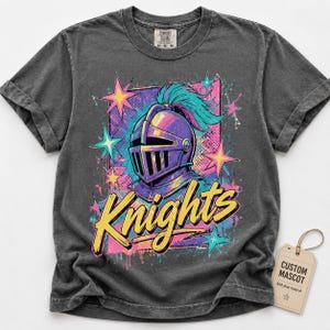 May include: A charcoal gray short-sleeve t-shirt featuring a graphic of a purple knight helmet with a teal plume. The design includes yellow text reading Knights, pink and teal stars, and a rectangular background. A beige tag reads CUSTOM MASCOT.