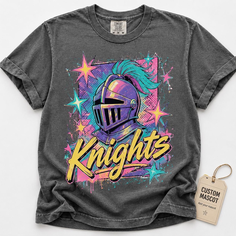 May include: A charcoal gray short-sleeve t-shirt featuring a graphic of a purple knight helmet with a teal plume. The design includes yellow text reading Knights, pink and teal stars, and a rectangular background. A beige tag reads CUSTOM MASCOT.