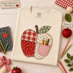 May include: A cream-colored crewneck t-shirt featuring a large apple graphic composed of various fabric patterns, including red plaid, red floral, and green floral prints. The apple includes a small pocket containing three colored pencils.
