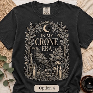 K&ouml;nnte beinhalten: Ein schwarzes Kurzarm-T-Shirt mit einem beigen Grafikdruck eines Raben, Pilzen, Kerzen und einer Mondsichel. Der Text lautet IN MY CRONE ERA. Das Design ist von einer bogenf&ouml;rmigen botanischen Umrandung mit Farnen und floralen Elementen umgeben.