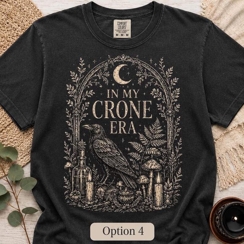 K&ouml;nnte beinhalten: Ein schwarzes Kurzarm-T-Shirt mit einem beigen Grafikdruck eines Raben, Pilzen, Kerzen und einer Mondsichel. Der Text lautet IN MY CRONE ERA. Das Design ist von einer bogenf&ouml;rmigen botanischen Umrandung mit Farnen und floralen Elementen umgeben.