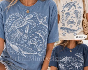 May include: Three short-sleeve t-shirts featuring white line art of marine life including sea turtles, whales, dolphins, jellyfish, crabs, and stingrays. Option 1 is blue, Option 2 is cream, and Option 3 is dark blue. Text reads UrbanMintDesignShop.