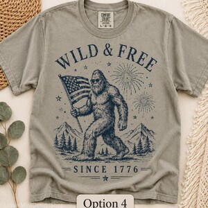 Puede incluir: Camiseta verde salvia con un gr&aacute;fico de Bigfoot sosteniendo una bandera estadounidense, fuegos artificiales y el texto "WILD & FREE SINCE 1776". La camiseta tiene cuello redondo y mangas cortas. La marca "COMFORT COLORS" est&aacute; impresa en el cuello.