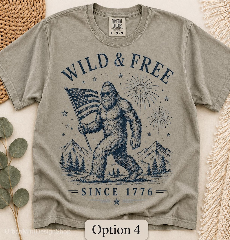 Puede incluir: Camiseta verde salvia con un gr&aacute;fico de Bigfoot sosteniendo una bandera estadounidense, fuegos artificiales y el texto "WILD & FREE SINCE 1776". La camiseta tiene cuello redondo y mangas cortas. La marca "COMFORT COLORS" est&aacute; impresa en el cuello.
