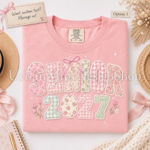 May include: A pink t-shirt featuring the word SENIOR and the year 2027 in a patchwork style with floral and gingham patterns. The design includes a pink bow above the text and small floral illustrations. The shirt has a Comfort Colors brand label.