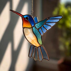 May include: Stained glass hummingbird with outstretched wings. The bird features a red beak, blue head, and wings in shades of blue and green. The body is a mix of orange and cream colors, with a gold-colored metal outline.