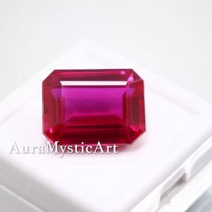 Op de afbeelding: Een rechthoekige, gefacetteerde, magenta edelsteen wordt gepresenteerd op een wit oppervlak. De edelsteen is geplaatst in een witte doos. De tekst "AuraMysticArt" is op de doos gedrukt.