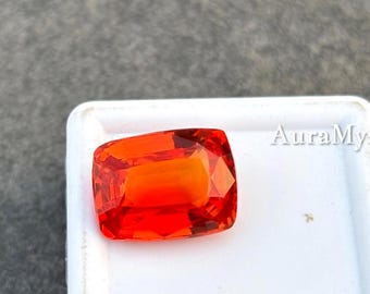 May include: A vibrant, rectangular cushion-cut gemstone with a brilliant orange hue and faceted surface, displayed inside a white plastic display case. The gemstone exhibits deep saturation and light reflection across its polished top facets.
