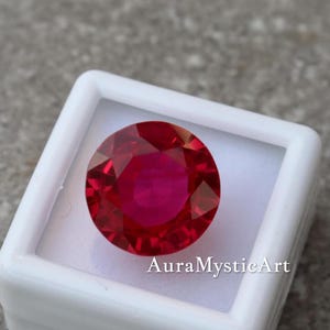 May include: A round, faceted ruby gemstone, presented in a white square box. The ruby is a deep red colour with a brilliant shine. The text "AuraMysticArt" is printed on the box.