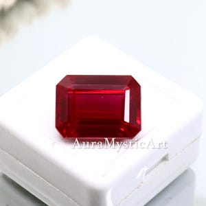 May include: A vibrant, deep red gemstone with a rectangular emerald cut, featuring sharp facets and high clarity. The stone is positioned on a white display box, with the text AuraMysticArt printed in a dark font across the lower portion.