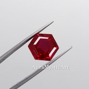 May include: A faceted, hexagonal ruby gemstone held by tweezers. The gem is a deep red color with a bright, reflective surface. The text "AuraMysticArt" is printed in white below the gemstone.