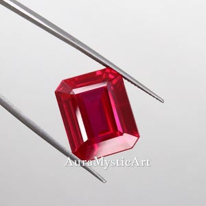 Puede incluir: Una gema de rubí rectangular de corte esmeralda sostenida por pinzas de metal plateado. La piedra muestra un tono rojo profundo y saturado con facetas claras que reflejan la luz. El texto AuraMysticArt es visible en color blanco en la parte inferior.