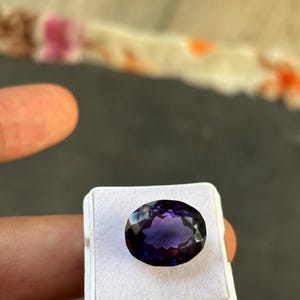 May include: An oval-cut, faceted gemstone with a deep purple hue, set in a white display case. The gem's surface reflects light, showcasing its multiple facets. The background is a blurred mix of colors.