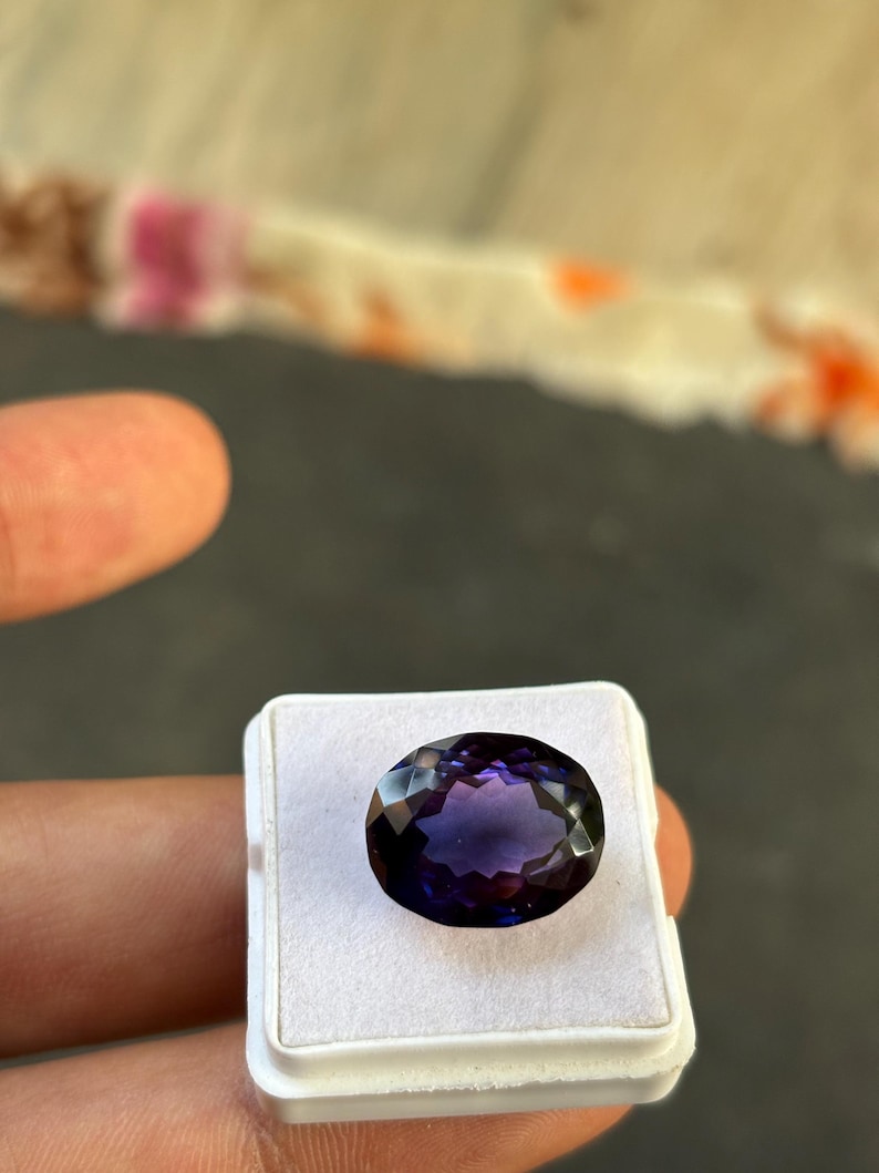 May include: An oval-cut, faceted gemstone with a deep purple hue, set in a white display case. The gem's surface reflects light, showcasing its multiple facets. The background is a blurred mix of colors.