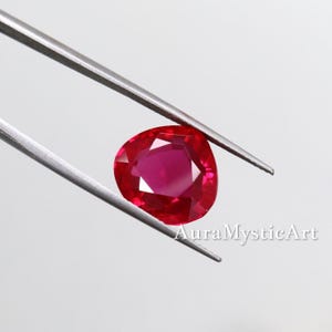 May include: A vibrant, heart-shaped ruby gemstone held by silver tweezers. The ruby displays a deep red hue with facets that catch the light. The text "AuraMysticArt" is visible in the lower right corner.