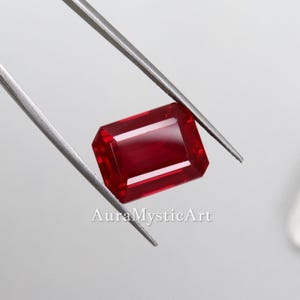 以下が含まれることがあります： 銀色の金属製ピンセットで保持された、長方形のファセットカットが施された深い赤色の宝石。この石は、鋭いエッジと磨かれた表面を持ち、鮮やかで透明感のある深紅の色合いを呈しています。宝石の下には、白い文字でAuraMysticArtと印字されています。