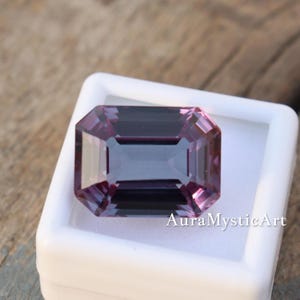 May include: A rectangular-cut gemstone in shades of purple, displayed in a white case. The gem's facets catch the light, showcasing its colour. The text "AuraMysticArt" is printed on the case.