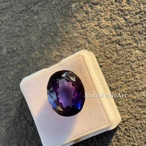 May include: An oval-cut amethyst gemstone with deep purple and blue hues, set in a white display case. The gemstone is faceted, reflecting light. The text "AuraMysticArt" is visible on the case.