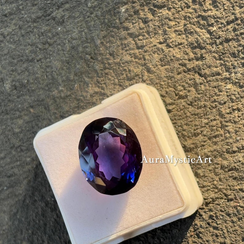 May include: An oval-cut amethyst gemstone with deep purple and blue hues, set in a white display case. The gemstone is faceted, reflecting light. The text "AuraMysticArt" is visible on the case.