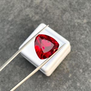 Op de afbeelding: Een levendige, dieprode edelsteen geslepen in een driehoekige vorm met meerdere facetten die het licht reflecteren. De edelsteen wordt vastgehouden door een dunne, zilverkleurige metalen pincet, gepositioneerd boven een klein, vierkant wit doosje.
