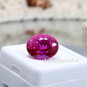 May include: A vibrant, oval-cut fuchsia gemstone with a faceted surface that reflects light, resting on a white plastic display box. The text AuraMysticArt is printed in a dark font on the front of the white box, positioned below the gemstone.