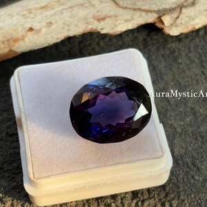 May include: An oval-cut gemstone with a deep purple and blue color gradient. The gem is set in a white plastic case, with the text "AuraMysticArt" visible on the right side. The gemstone is faceted, reflecting light.