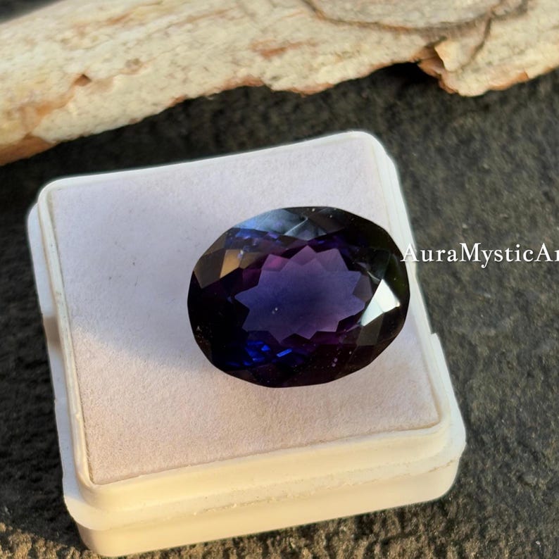 May include: An oval-cut gemstone with a deep purple and blue color gradient. The gem is set in a white plastic case, with the text "AuraMysticArt" visible on the right side. The gemstone is faceted, reflecting light.