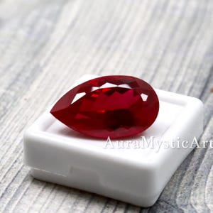 Op de afbeelding: Een peervormige, gefacetteerde rode edelsteen die op een wit vierkant doosje rust. De edelsteen heeft een diepe, levendige rode kleur met meerdere geometrische facetten die het licht reflecteren voor een schitterend effect. De tekst AuraMysticArt is zichtbaar.