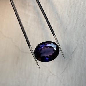 May include: An oval-cut, deep purple gemstone is held by a pair of tweezers. The gem has multiple facets that reflect light, creating a sparkling effect. The tweezers are silver-colored and have sharp, pointed tips.