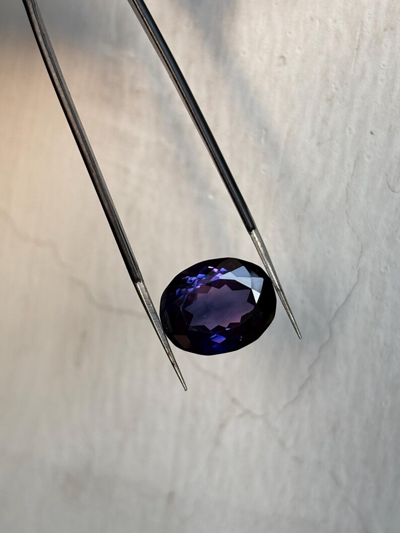 May include: An oval-cut, deep purple gemstone is held by a pair of tweezers. The gem has multiple facets that reflect light, creating a sparkling effect. The tweezers are silver-colored and have sharp, pointed tips.