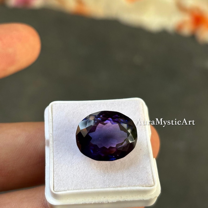 May include: An oval-cut, faceted gemstone with deep purple and violet hues. The gem is set in a white display case, with the text "AuraMysticArt" visible on the right side. The gemstone is held by a person.