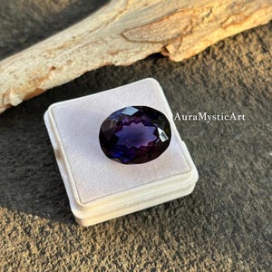 May include: An oval-cut, deep purple gemstone with facets, set in a white display box. The gem has a dark, lustrous appearance, with hints of blue and violet. The text "AuraMysticArt" is visible on the right side.