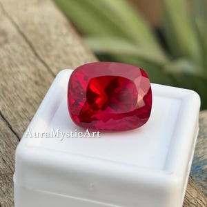 Puede incluir: Una gema roja vibrante de corte cojín con una superficie facetada que refleja la luz, exhibida sobre un recipiente cuadrado de plástico blanco. El texto AuraMysticArt está impreso en blanco en la parte inferior izquierda del recipiente.