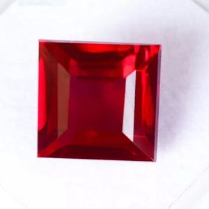 May include: A square-cut, faceted gemstone in a deep, vibrant red hue. The gemstone has a polished surface with sharp, clean edges, reflecting light. The gem is set against a white background, highlighting its colour and clarity. This gemstone is suitable for jewellery.