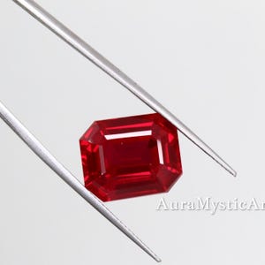 Könnte beinhalten: Ein facettierter, rechteckig geschliffener Rubin, der von einer silbernen Pinzette gehalten wird. Der Rubin hat eine tiefe, leuchtend rote Farbe mit scharfen Kanten und einer polierten Oberfläche. Der Hintergrund ist weiß.