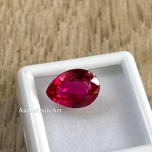 May include: A pear-shaped, faceted ruby gemstone in a white display box. The ruby is a deep, vibrant red with a polished surface. The text "AuraMysticArt" is printed on the lower left side of the box.