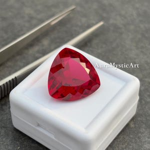 Può includere: Una gemma rosso scuro sfaccettata, di forma triangolare. La gemma è posta in un astuccio di plastica bianco, con delle pinzette sullo sfondo. Il testo "AuraMysticArt" è visibile sul lato destro dell'immagine.