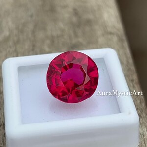 Puede incluir: Una gema de rubí redonda y facetada, de color rojo intenso, presentada en una caja blanca. El texto "AuraMysticArt" es visible en la caja. El fondo es una superficie de color marrón claro.
